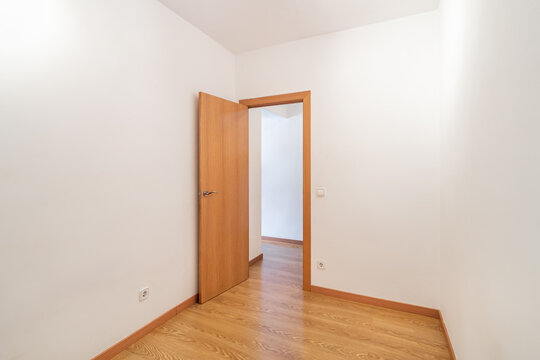 Empty room with wooden floor and white walls. A wooden door is open, leading to another room. Natural light illuminates the space.