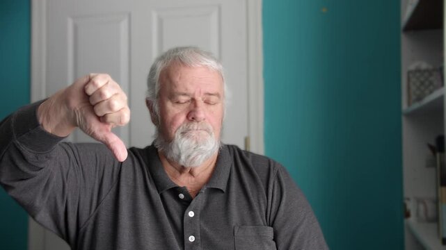 A senior person with gray hair is sitting in a room. They show a thumbs down with their hand while making a serious expression. This happens in a home setting.