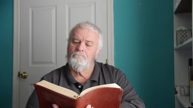 A man sits at a table and reads a book in a room. The light shines in through a window. He appears focused on the pages. The room has simple decor and a door in the background.