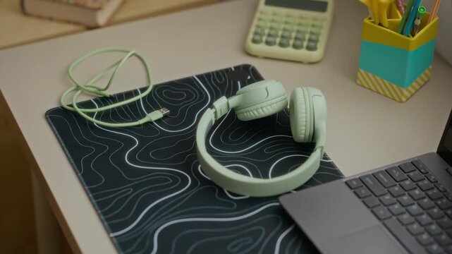 Green wireless headphones resting on mousepad next to laptop on desk in kid bedroom. Footage ideal for remote education, online learning, and modern workspace technology marketing campaigns