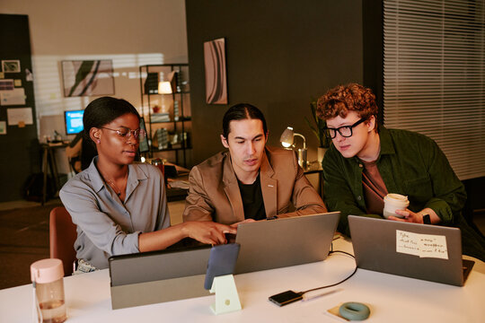 Medium shot of diverse coworkers reviewing laptop data, discussing project strategy during late office collaboration. Ideal for teamwork, startup, productivity marketing