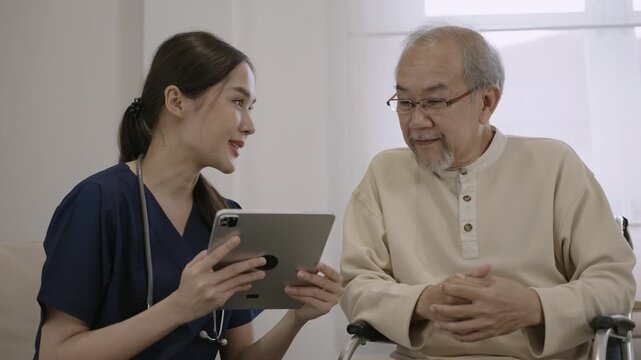 Professional young Asian woman nurse caregiver using a digital tablet to talk and provide advice about healthcare to a senior man patient while sitting in a wheelchair in a special room at hospital. 