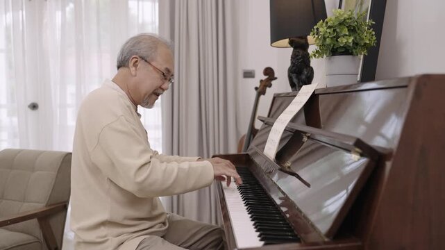 Asian senior man pianist in eyeglasses is sitting playing classical piano alone after retirement for relaxing in living room at home. Elderly male playing a keyboard organ instrument with happiness. 