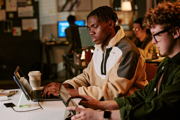 Obraz premium Medium shot of Black young adult man working on laptop beside Caucasian young adult man using tablet in shared office. Supporting teamwork, startup productivity, coworking marketing