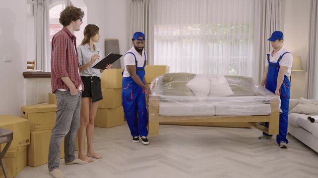 Two professional movers in uniform are moving furniture and cardboard boxes during a home relocation, while a young couple of homeowners, holding a clipboard, are checking for delivery at their home. 