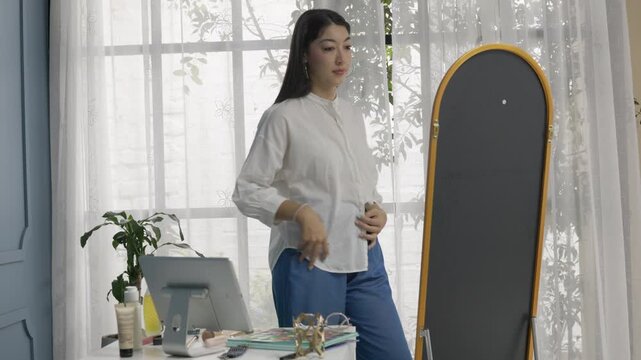 Young woman adjusting high waist pants while getting dressed in front of mirror at home