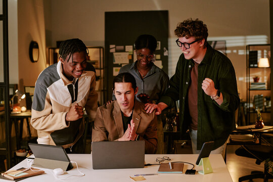 Diverse team celebrating success around laptop in office, collaborating on project results and startup growth. Ideal for business teamwork, innovation, leadership campaigns