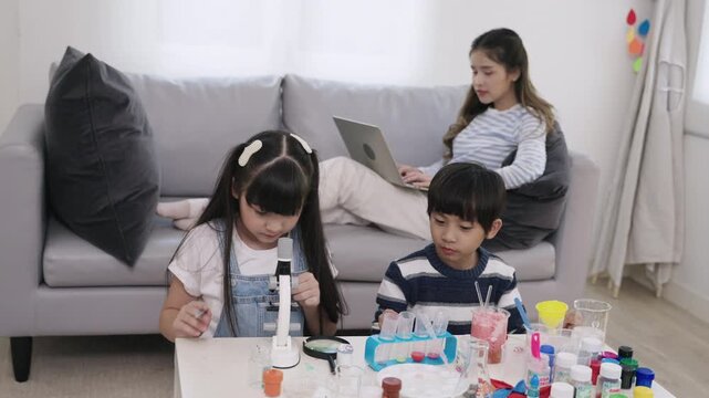 Happy adorable Asian boy and girl are sitting learning science and conducting chemistry experiments together with a young teacher sitting and taking care in the back of a nursery classroom lab. 