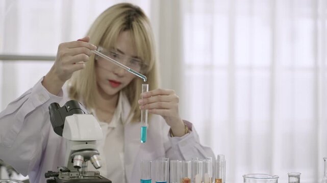 A professional Asian woman scientist and researcher in a lab coat wearing safety goggles with microscope is conducting chemistry experiments and mixing liquids in test tubes in the science laboratory.
