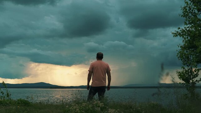 A lone man stands on the shore of a lake against a cloudy, stormy sky. Human psychology, time alone with oneself.