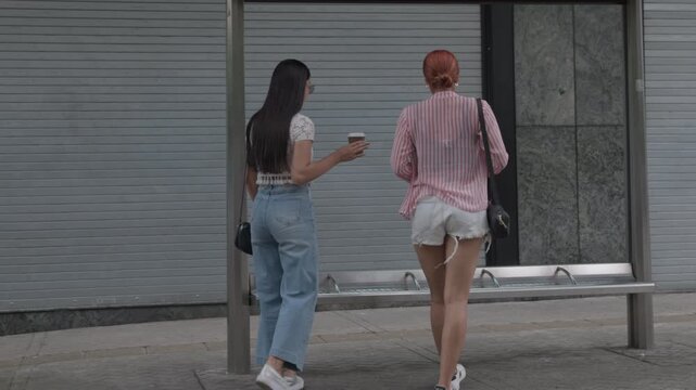 Two young women sitting at bus stop talking while drinking coffee