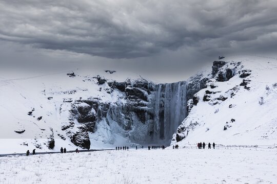 Tourists at Skogafoss waterfall, snowy and icy rock face, dark clouds above, Sudurland, Iceland
