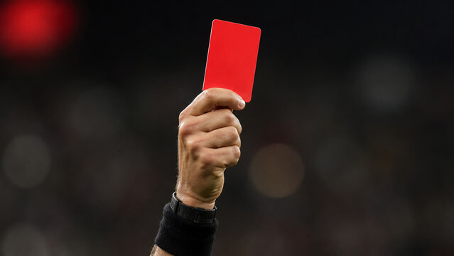 A referee's hand holding up a red card during a sports event, signaling a penalty or dismissal.