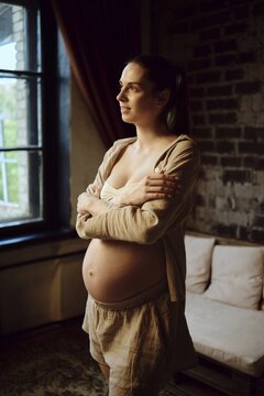 Pregnant woman standing near window at home, reflecting in quiet moment during evening