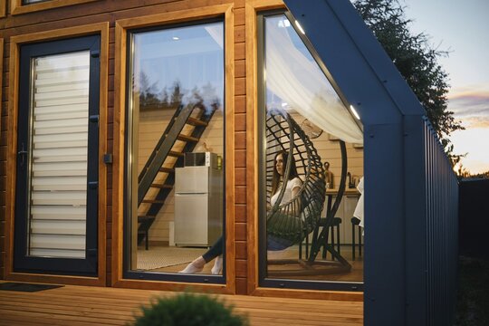 A cozy and stylish tiny house interior captured during twilight. The image showcases a woman lounging comfortably in a unique egg chair, reflecting a serene lifestyle