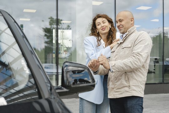 A couple is engaged in conversation while exploring a car at a dealership. The woman gestures playfully, suggesting enthusiasm, while the man examines the vehicle's details