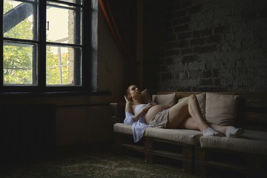 A pregnant woman lounges comfortably on a cozy couch in her home. The warm sunlight filters through the window, creating a serene atmosphere