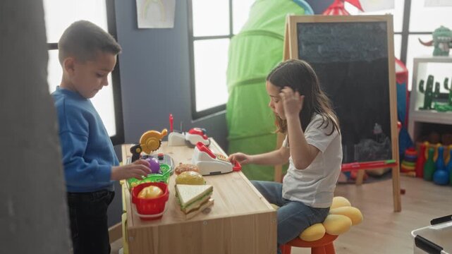 Girl uses toy cash register with hand pressing buttons while boy hands her play fruit in a building; playful cooperation.