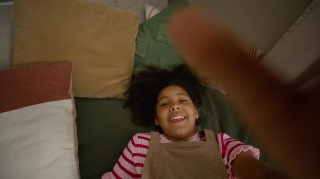 Portrait of African American child looking at camera, smiling, and falling backward onto bed while filming selfie video. Footage suiting youth lifestyle vlogs or family marketing campaigns