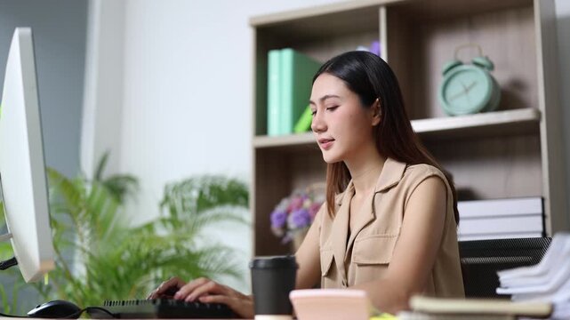 Young Asian businesswoman working on computer, typing on keyboard and smiling, sitting at her desk in a modern office, enjoying her job and looking happy and focused
