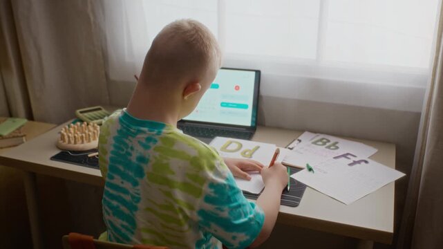 Back view of boy with Down syndrome studying at desk, writing alphabet letters on paper worksheet and using laptop. Footage perfect for inclusive education and special needs learning promos