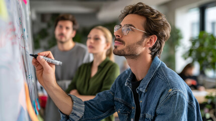 Fototapeta premium Young man drawing with marker on whiteboard, colleagues watching, creative teamwork, focused mood