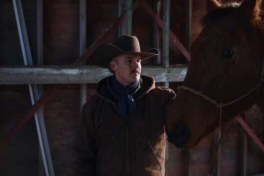 Cowboy interacts with horse inside barn during daylight hours