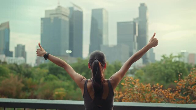 Back view of happy Asian woman in sportswear raising arms and showing thumbs up in urban green park, successful female athlete celebrating workout achievement with city skyscraper, fitness and health