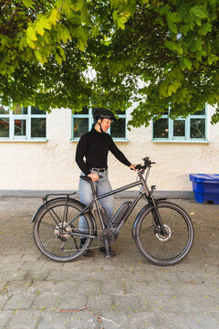 Female cyclist in helmet standing with electric bike under trees