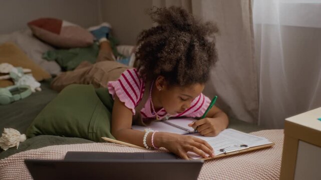 African American girl lying on bed, writing on clipboard and using laptop for schoolwork. Ideal for commercial videos promoting online education platforms and remote learning technology