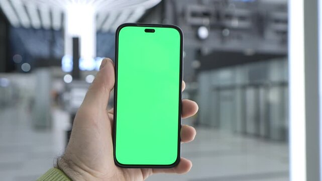 Hand holds smartphone with green screen. Background shows blurred modern indoor space. Three keyframes show same scene. Hand wears light green sleeve. Phone has black frame and notch