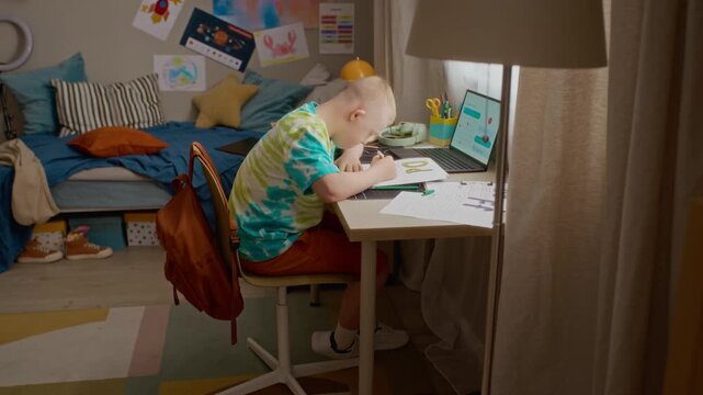 Side view of boy with Down syndrome studying at desk, writing on paper and using laptop for online school. Footage representing inclusive education and special needs student support