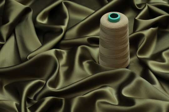 Luxurious 19 momme dark olive green silk satin fabric, showcasing a soft, flowy drape with thread atop. Perfect for high-end fashion, sophisticated evening wear and premium bedding.