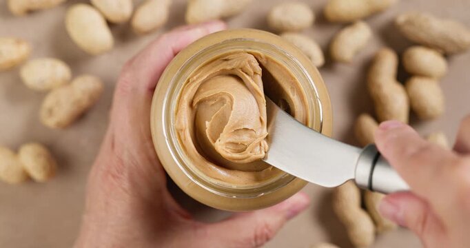 human hand with a spreadable kitchen knife takes out creamy peanut butter from a jar to prepare a healthy breakfast