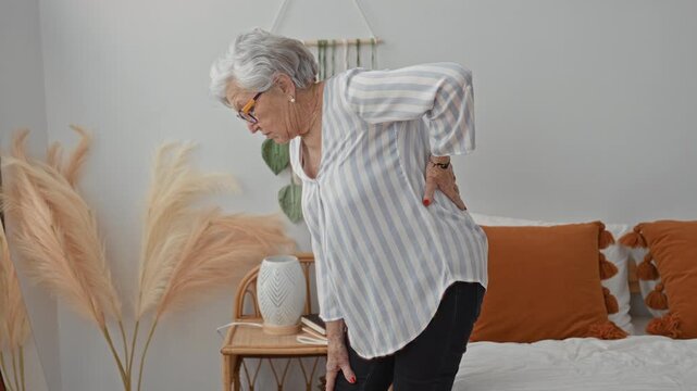 Woman holding lower back and bending beside bed in a bedroom of a residential building; discomfort aging.