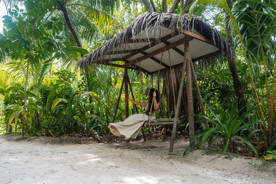 A woman in a flowing white dress sits on a wooden swing under a thatched palm roof at a Maldives resort, with sandy ground, palm trees, soft daylight, and a secluded mood.
