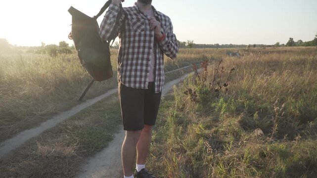 A cyclist in a helmet rolls down and closes his black roll-top backpack, puts it on, and walks out of the frame to continue his bicycle journey along a field trail.