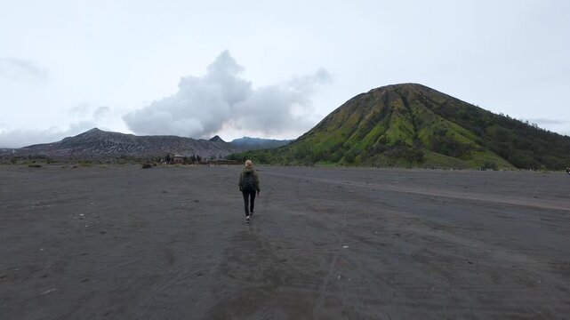FPV drone shot of a traveler crossing the Sea of Sand with Mount Batok rising in the background near Mount Bromo, East Java, Indonesia. Dramatic volcanic landscape and adventure travel.