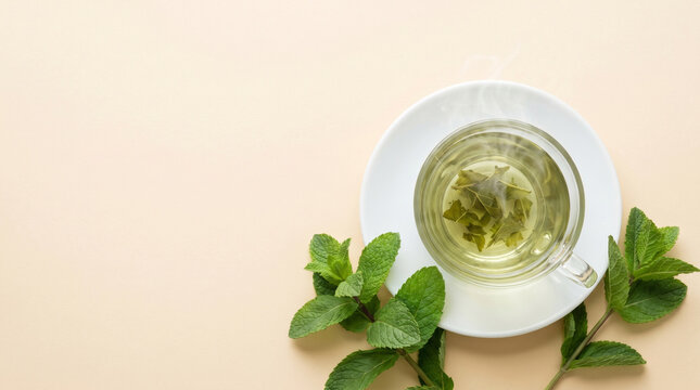 Top view of a steaming cup of fresh peppermint tea with green mint leaves on a light beige background