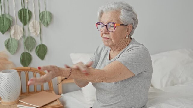 Woman massaging hands and rubbing wrists while seated on bed in a bright bedroom, wearing glasses and pearl earrings with red nails; self care relief.
