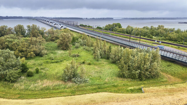 Aerial view of Moerdijk railway bridges spanning the broad Hollands Diep river, surrounded by lush green vegetation and trees under an overcast sky in Moerdijk, North Brabant, Netherlands.