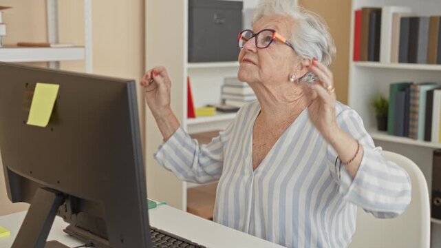 Woman at computer desk wearing glasses and striped blouse, hands raised with fingers poised near monitor in building; frustration resolve.