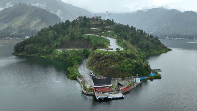 Aerial view of the winding Sibea Bea road and statue on a lush green hill surrounded by the waters of Lake Toba in Samosir Regency, North Sumatra, Indonesia.