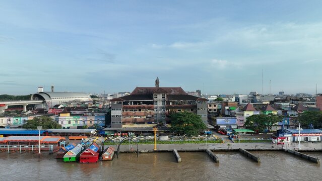 Aerial view of the Musi River waterfront featuring traditional boats, colorful buildings, and the iconic Ampera Bridge in Kecamatan Seberang Ulu I, Sumatera Selatan, Indonesia.