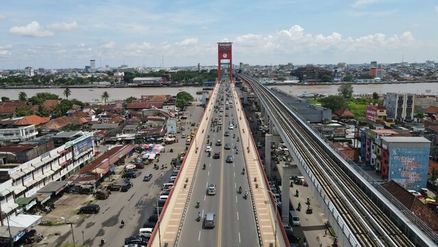 Aerial view of Jembatan Ampera crossing the Musi River with traffic, adjacent light rail tracks, and surrounding urban buildings in Palembang, Sumatera Selatan, Indonesia.