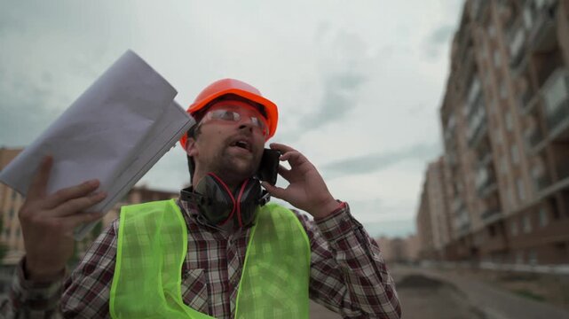 Low angle shot of a furious builder or engineer talking on a smartphone, showing an emotional reaction and holding construction plans against a cloudy sky background.