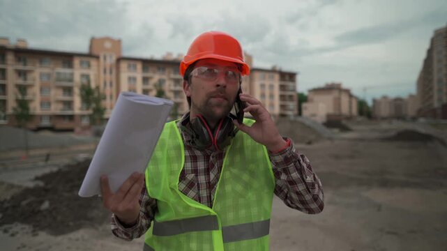 An angry construction foreman in an orange hardhat and safety vest argues on the phone while emotionally waving architectural blueprints on a residential building site.