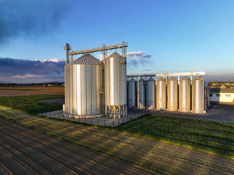 Aerial view of a row of metallic grain silos situated in a rural agricultural field during the golden hour in Villafranca Piemonte, Piedmont, Italy.