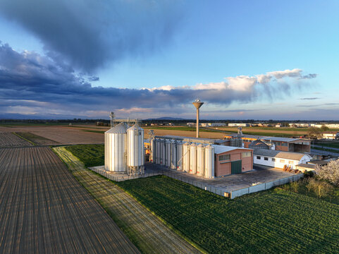Aerial view of a grain silo facility surrounded by agricultural fields under a dramatic cloudy sky in Villafranca Piemonte, Piedmont, Italy.