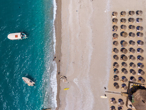 Aerial view of a sandy beach with rows of straw umbrellas, turquoise sea, and small boats in Sarande, Vlore County, Albania.